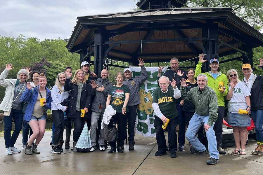 Large group of Baylor alumni gathered outdoors near a gazebo, raising their hands and showing sic 'em signs at a Dr Pepper Hour Tour event