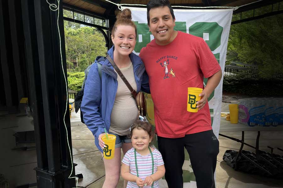 Baylor alumni couple and their young child posing in front of a Baylor banner at a Dr Pepper Hour Tour event, holding BU cups