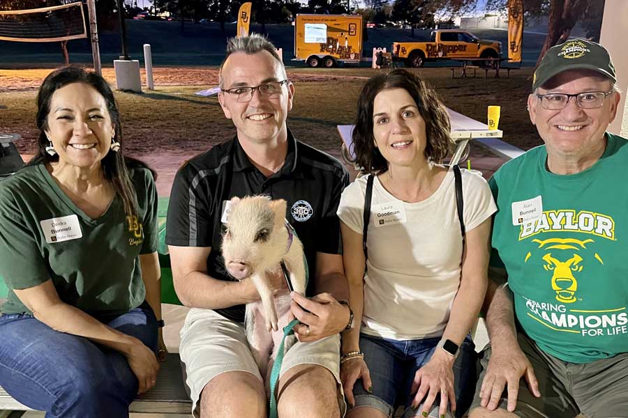 Four Baylor alumni seated at an outdoor nighttime event, one holding a small white piglet, with the Dr Pepper Hour Tour truck visible in the background