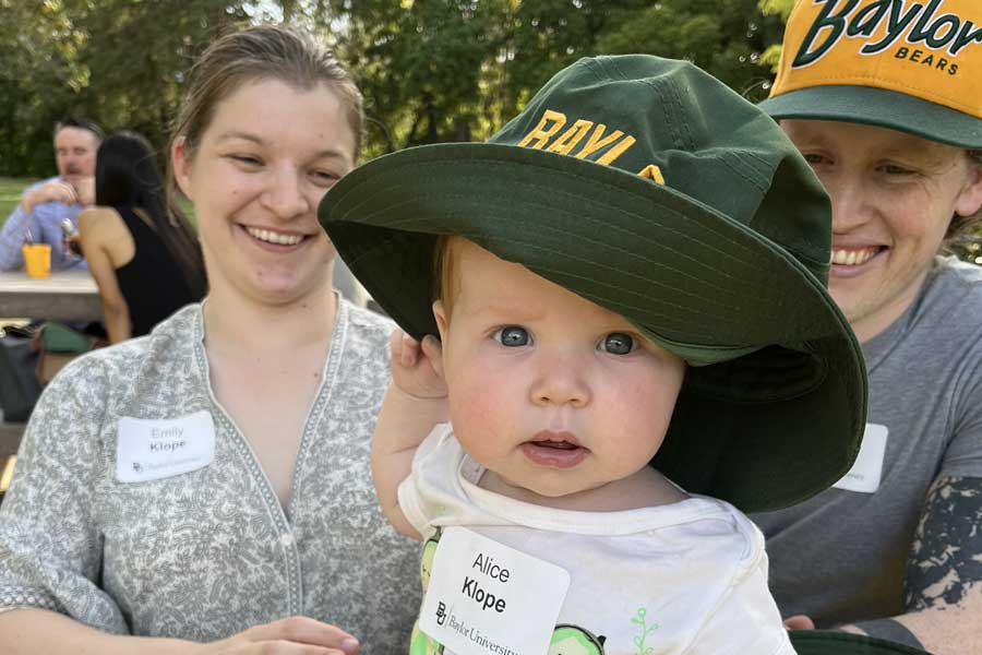 Baby wearing an oversized Baylor green hat, with two smiling parents at a Baylor alumni event