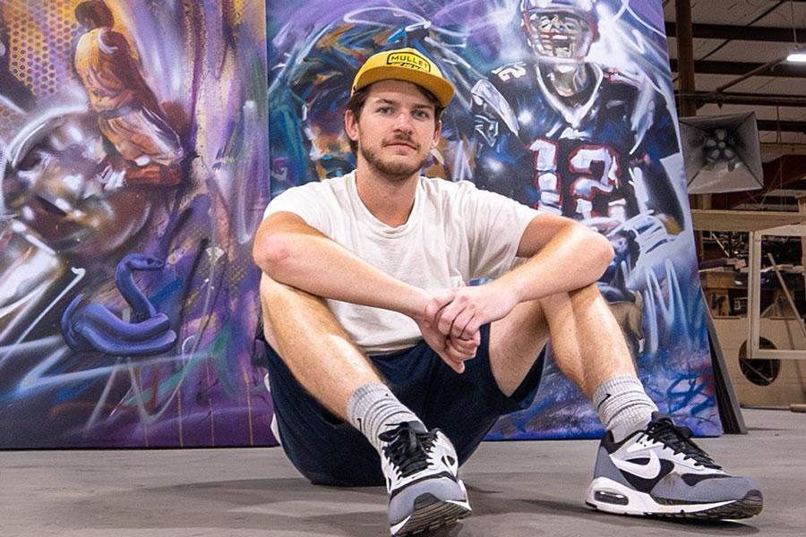 Artist Cade Kegerreis, seated in front of commission portraits of Kobe Bryant and Tom Brady