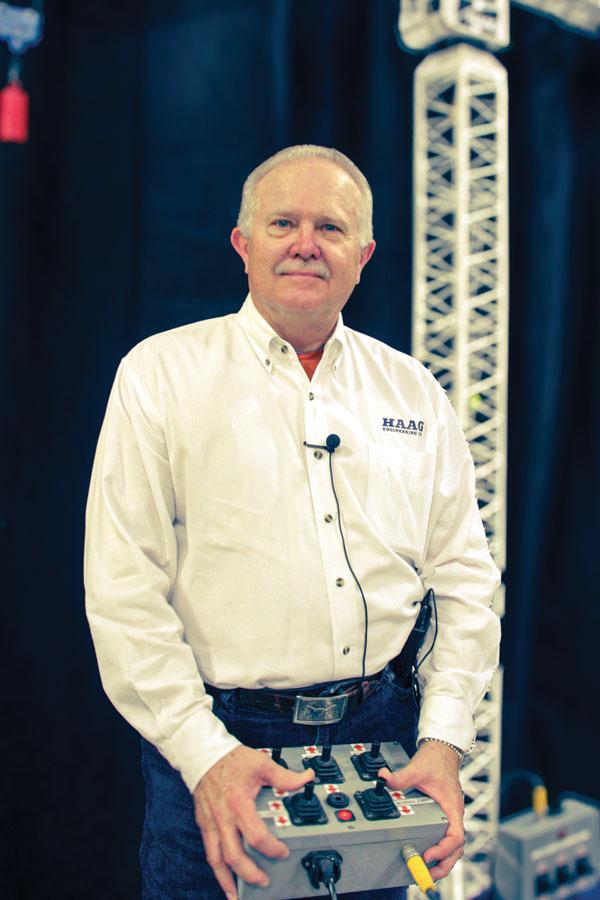 Jim Wiethorn holds a crane remote control in front of a lattice crane tower, wearing a white Haag Engineering shirt.