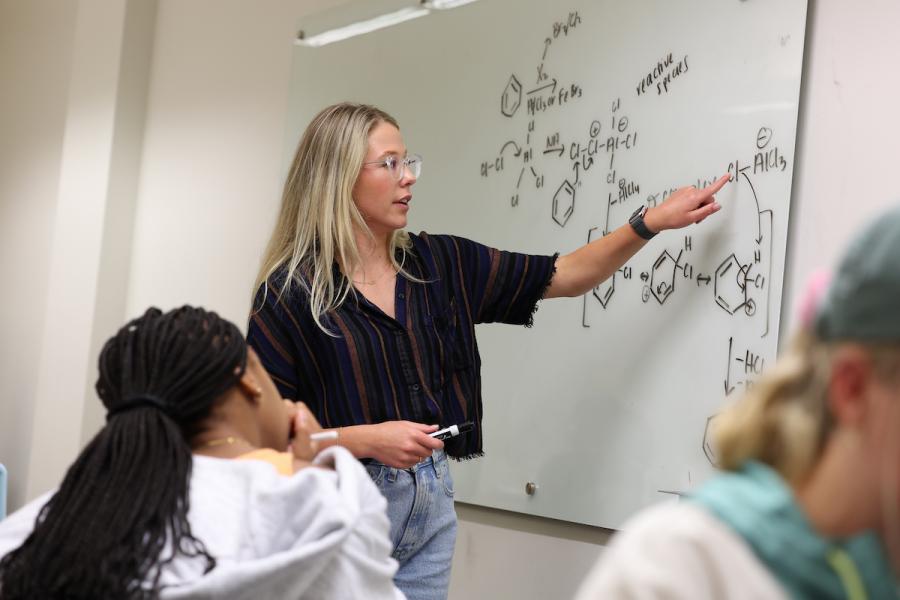A blonde white female student writes a chemical equation on a dry erase board and discusses it with a black female student.